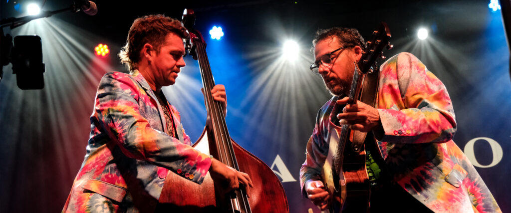 Andy Falco and Travis Book wearing tie-dyed suits under stage lights playing upright bass and acoustic guitar