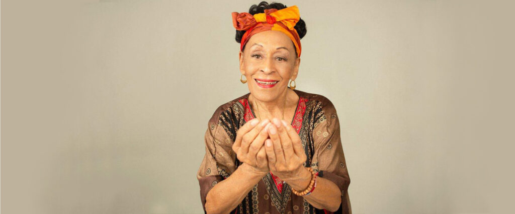 Omara Portuondo smiling with hands outstretched, palms cupped, wearing orange, red, and brown